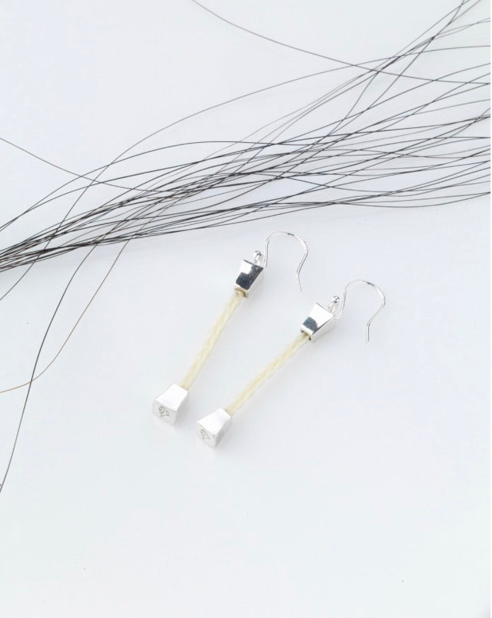 Forged Farriers Nail cream braided horse hair drop earrings in sterling silver, arranged beside loose black horse tail hairs.
